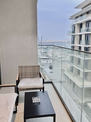 A balcony with a wicker chair and a small table, overlooking a marina with boats and buildings.
