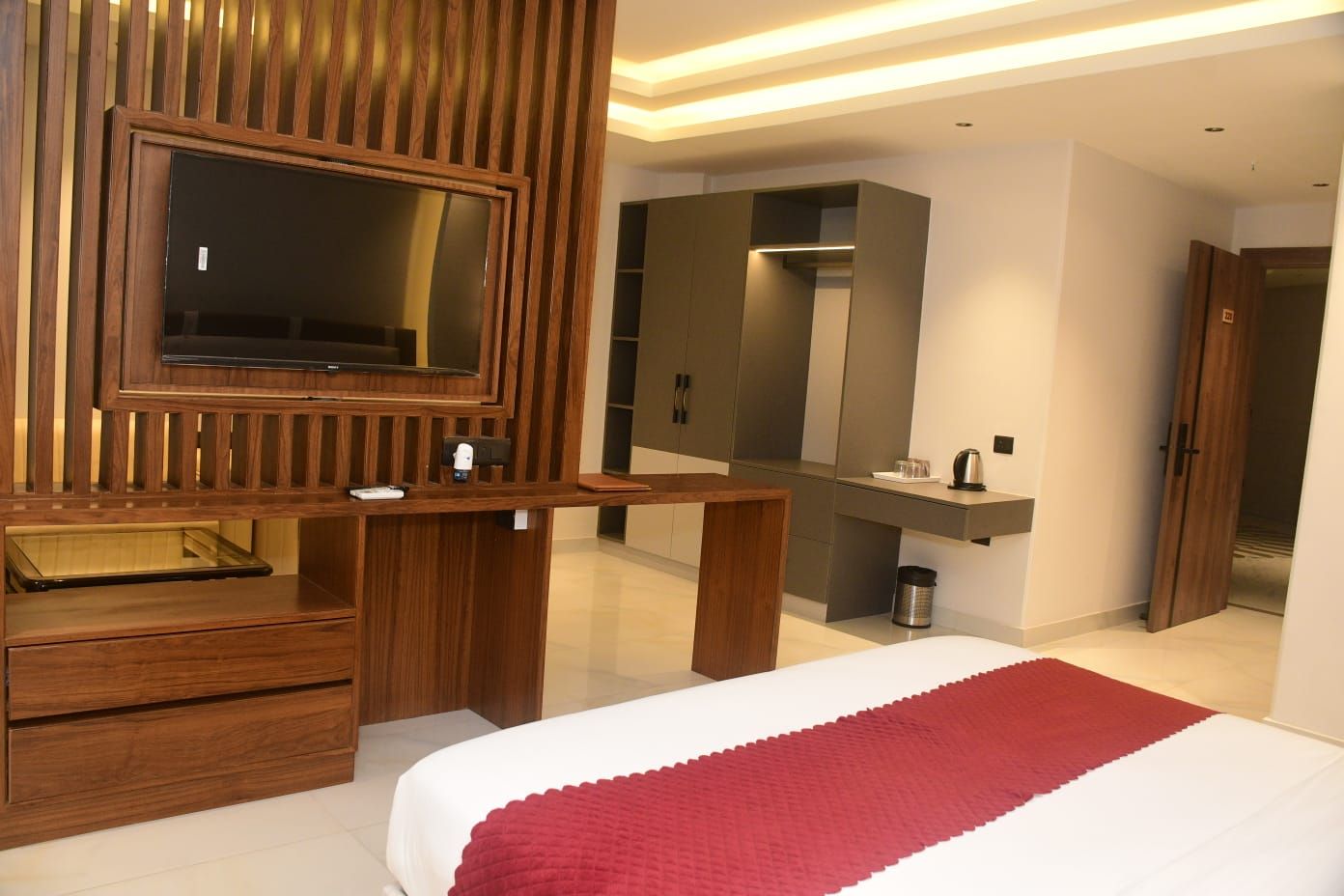 Habitación de hotel con un escritorio de madera, televisor, espejo grande, cama con manta roja y armario.