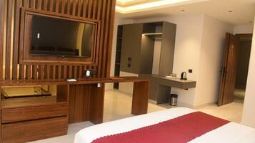 Habitación de hotel con un escritorio de madera, televisor, espejo grande, cama con manta roja y armario.