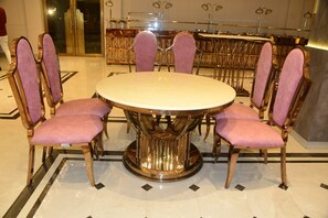 A round dining table with six chairs, each with a cushioned seat and backrest. The table has a metallic base and is surrounded by a tiled floor with a geometric pattern.