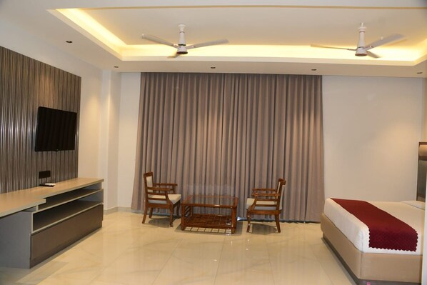 A modern hotel room with a flat-screen TV, a bed with a red throw, two wooden chairs, a small table, and a ceiling fan.