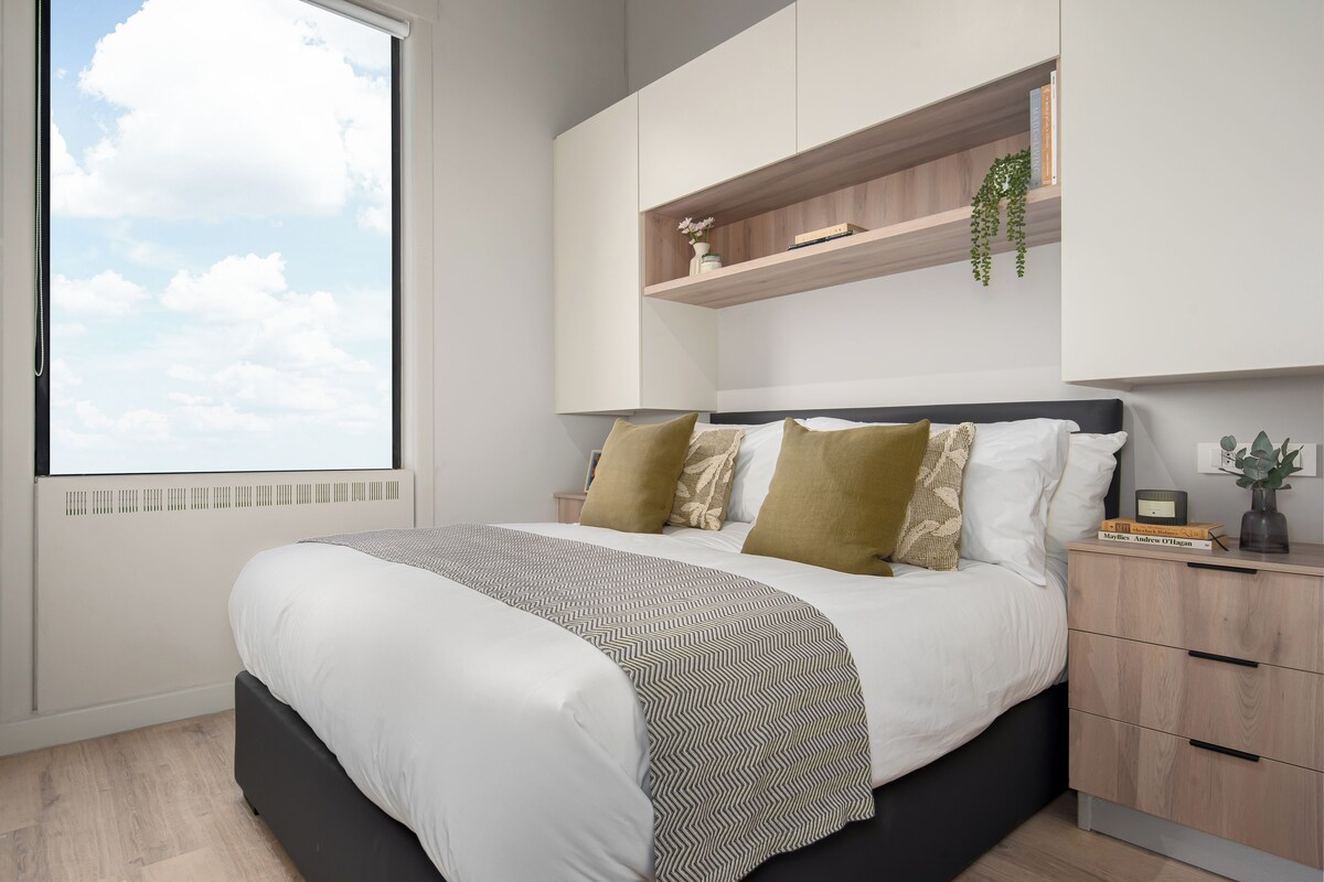 A modern bedroom with a large bed, a built-in wardrobe, and a window with a view of the sky.