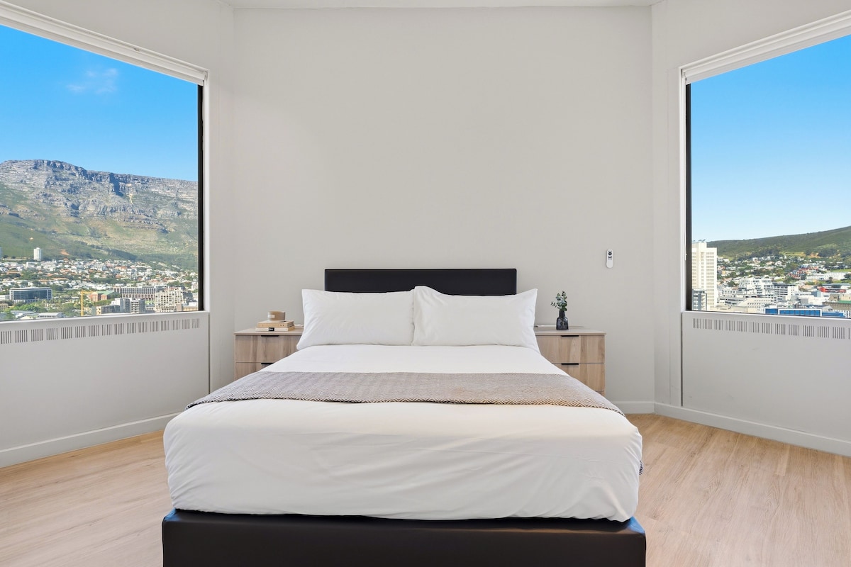 A bedroom with a bed, a nightstand, and a view of a cityscape through large windows.