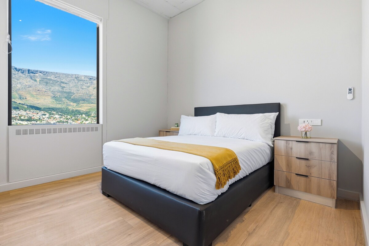 A bedroom with a bed, a nightstand, a bedside table, and a view of a mountainous landscape through a window.