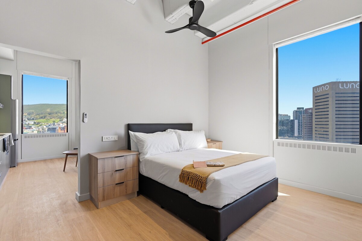 A modern bedroom with a bed, bedside tables, a ceiling fan, and a window with a city view.