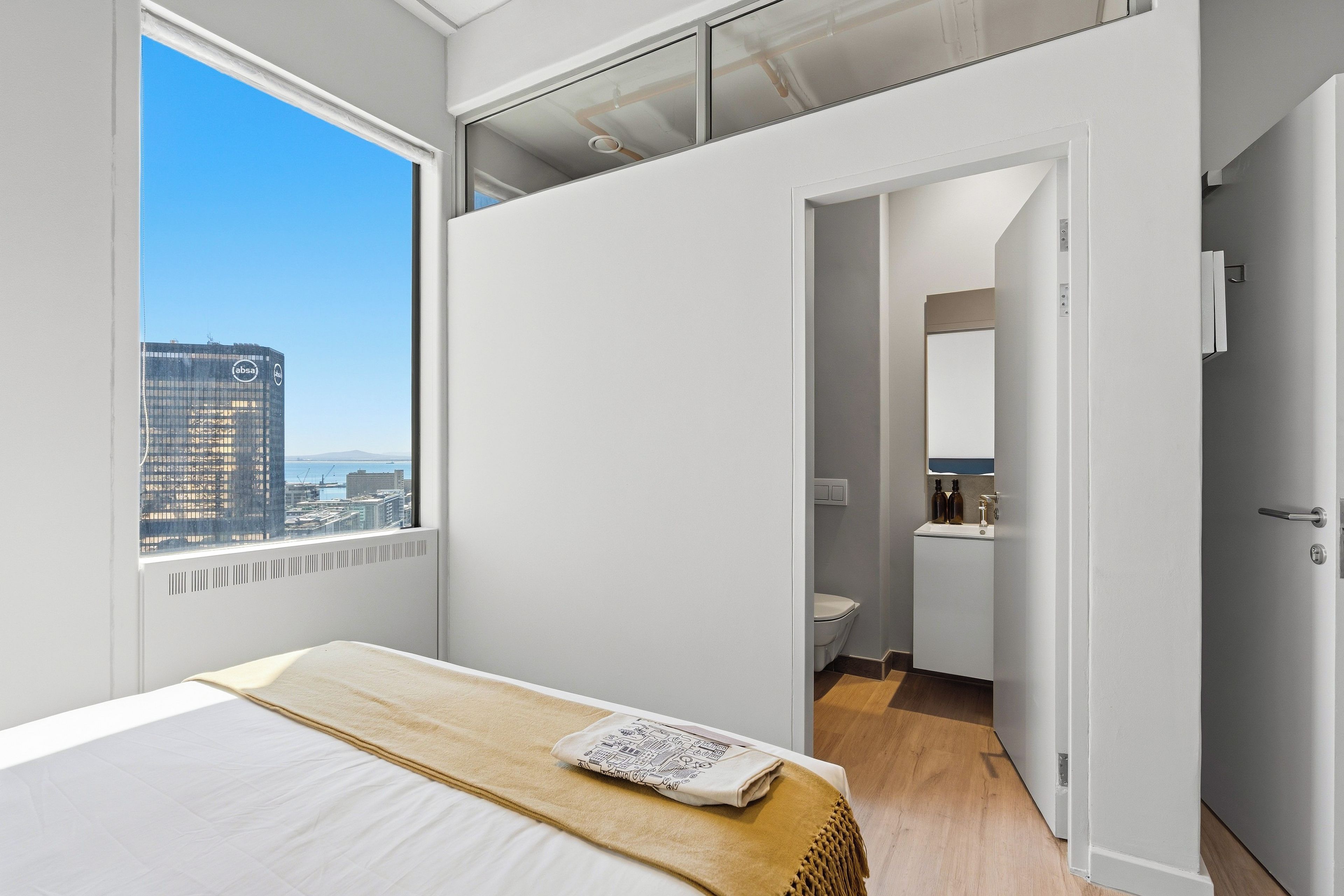 Habitación de hotel moderna con un ventanal grande, una cama con manta color beige, un baño pequeño y vista a la ciudad.
