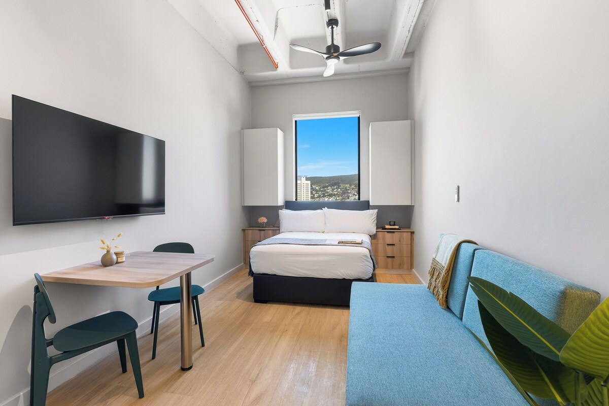 A compact bedroom with a bed, a TV, a desk, and a window.