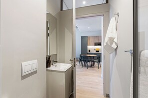 A modern bathroom with a sink and mirror, a hallway leading to a dining area with a table and chairs, and a towel hanging on a rack. - The Acre by Neighbourgood (Cape Town)
