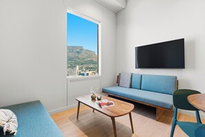 A modern living room with a sofa, a coffee table, a TV mounted on the wall, and a view of a cityscape through a large window. - The Acre by Neighbourgood (Cape Town)