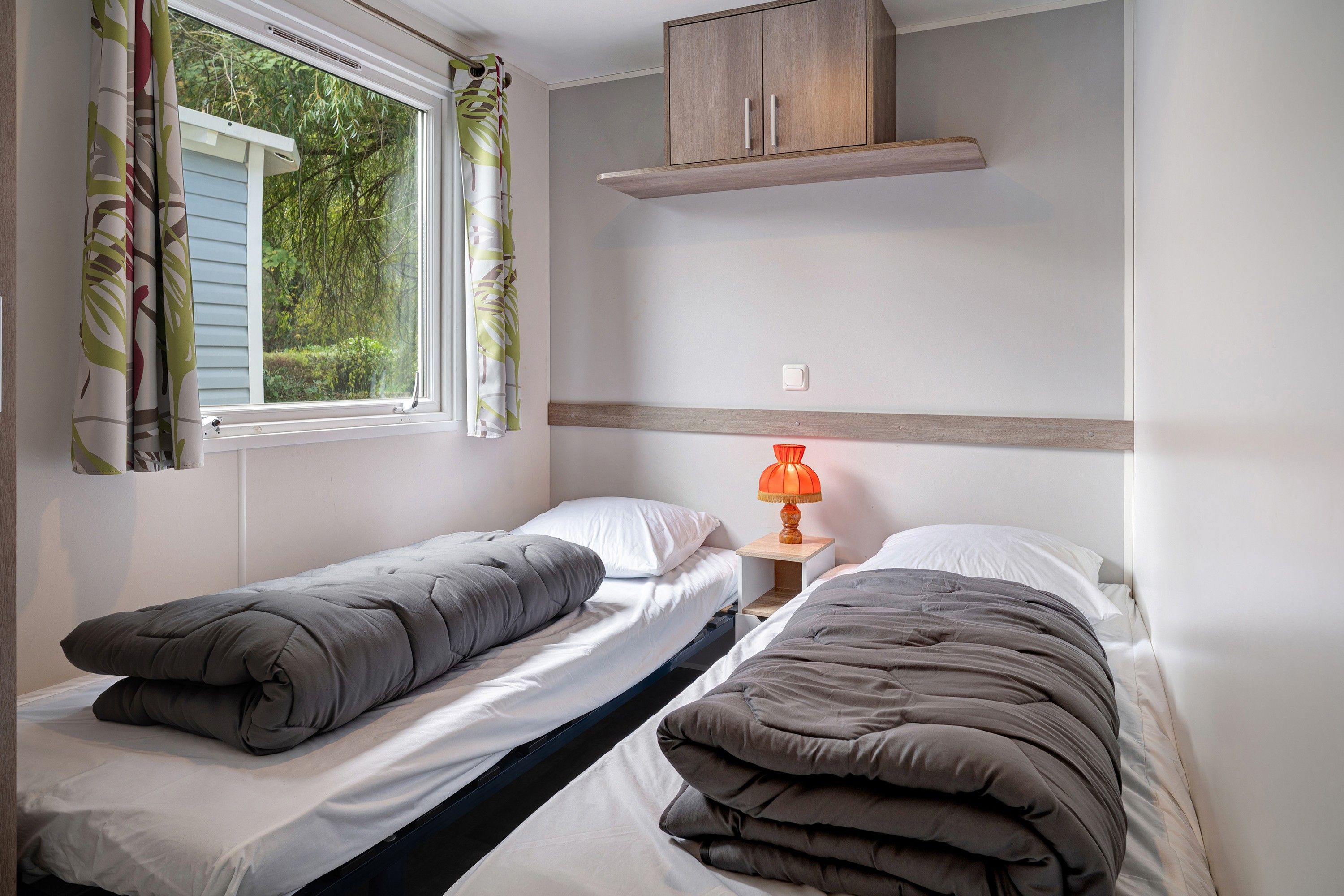 Un alojamiento pequeño, con una habitación individual, dos camas individuales, una ventana con cortinas, un estante de madera sobre las camas y una mesita de noche con lámpara.