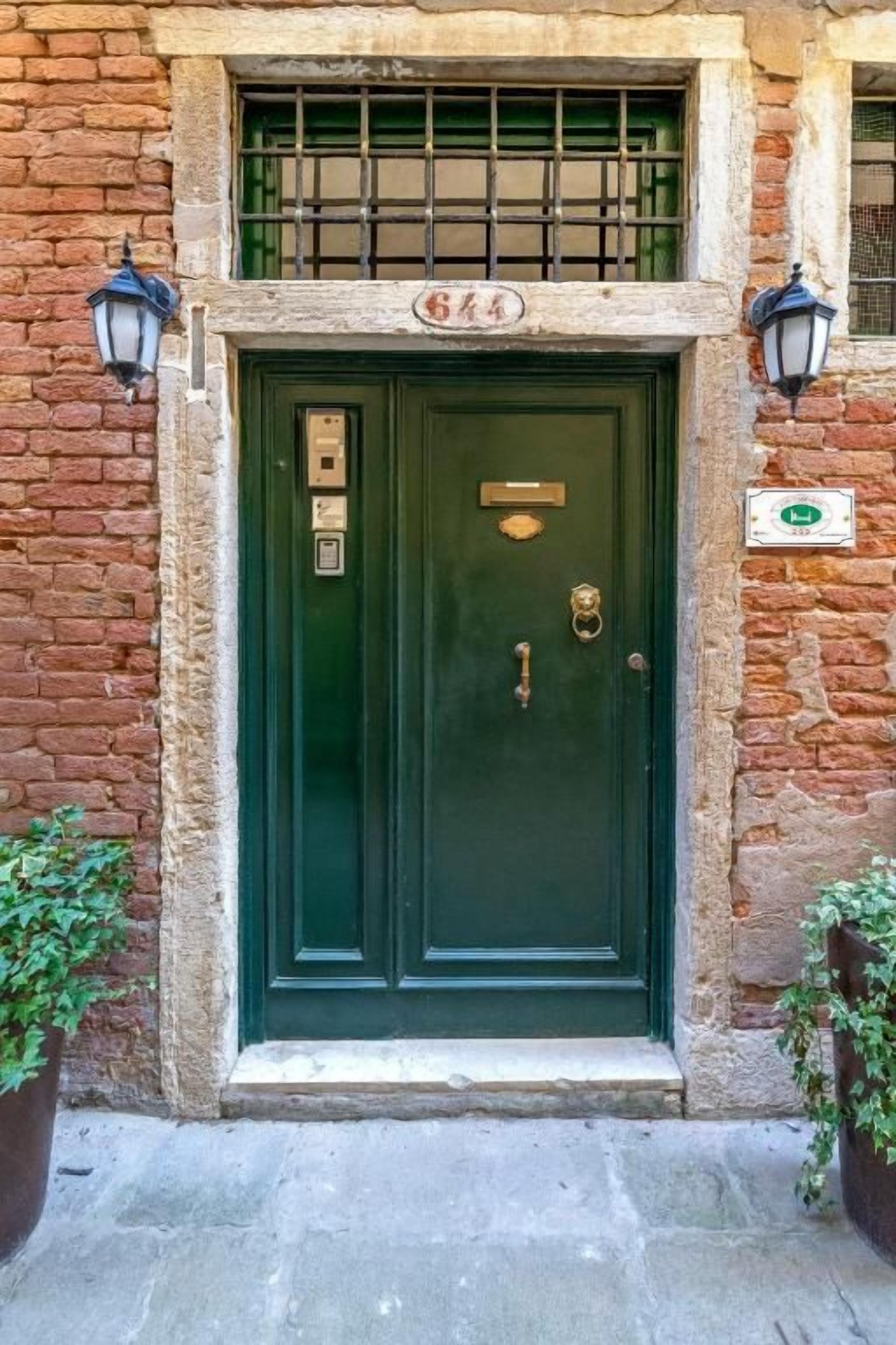 Une porte à deux battants, peinte en vert, avec une poignée en laiton, un heurtoir en laiton et une petite plaque en laiton au-dessus de la porte. Une fenêtre avec une grille métallique se trouve au-dessus de la porte, et deux lanternes sont fixées aux murs de part et d’autre.