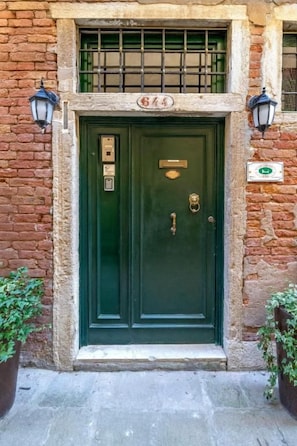 A green double door with a brass handle, a brass knocker, and a small brass plate above the door. There is a window with a metal grid above the door, and two wall-mounted lanterns on either side. - Casa Guggenheim (Venice)