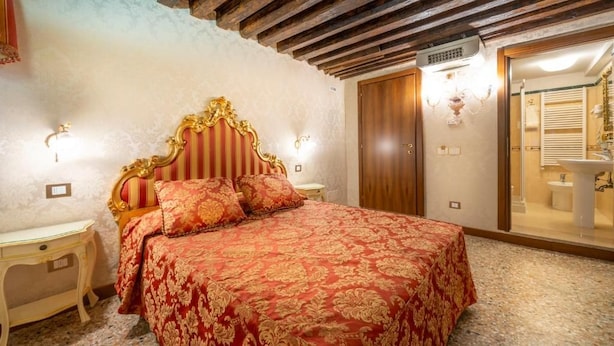 Ein Schlafzimmer mit einem großen Bett, Holzbalkendecke, einem Kleiderschrank und einem durch eine offene Tür einsehbaren Badezimmer.