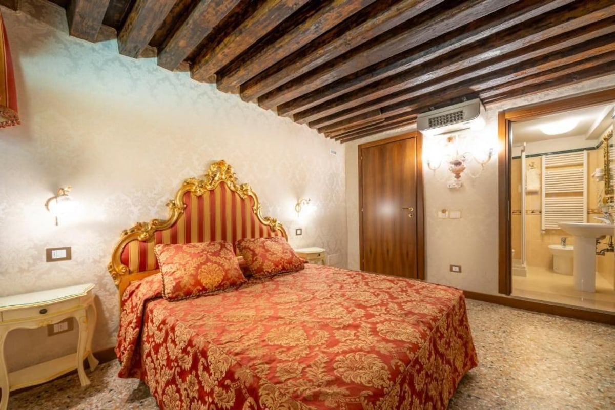 A bedroom with a large bed, wooden ceiling beams, a wardrobe, and a bathroom visible through an open door.