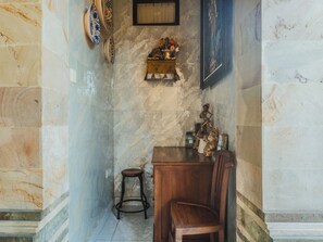 Un pasillo estrecho con un muro de mármol, un taburete de madera, una mesa de madera con una silla y una estantería con adornos.
