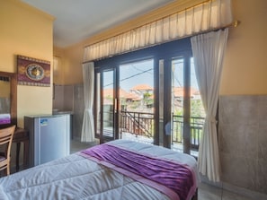 A hotel room with a bed, a desk, a refrigerator, and a balcony with a view of the neighborhood.