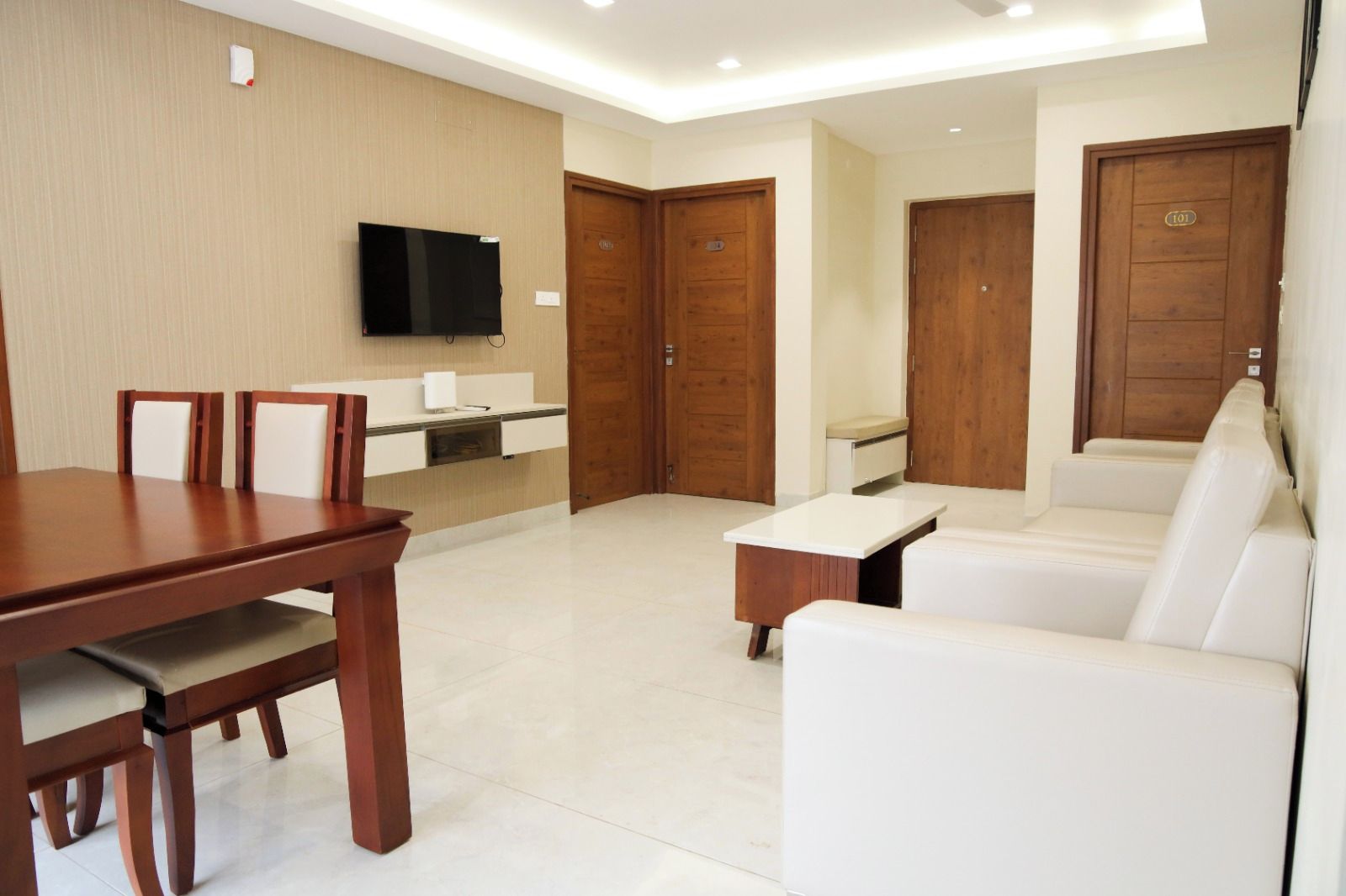 A modern living room with a dining table, white sofa, and a TV mounted on the wall.