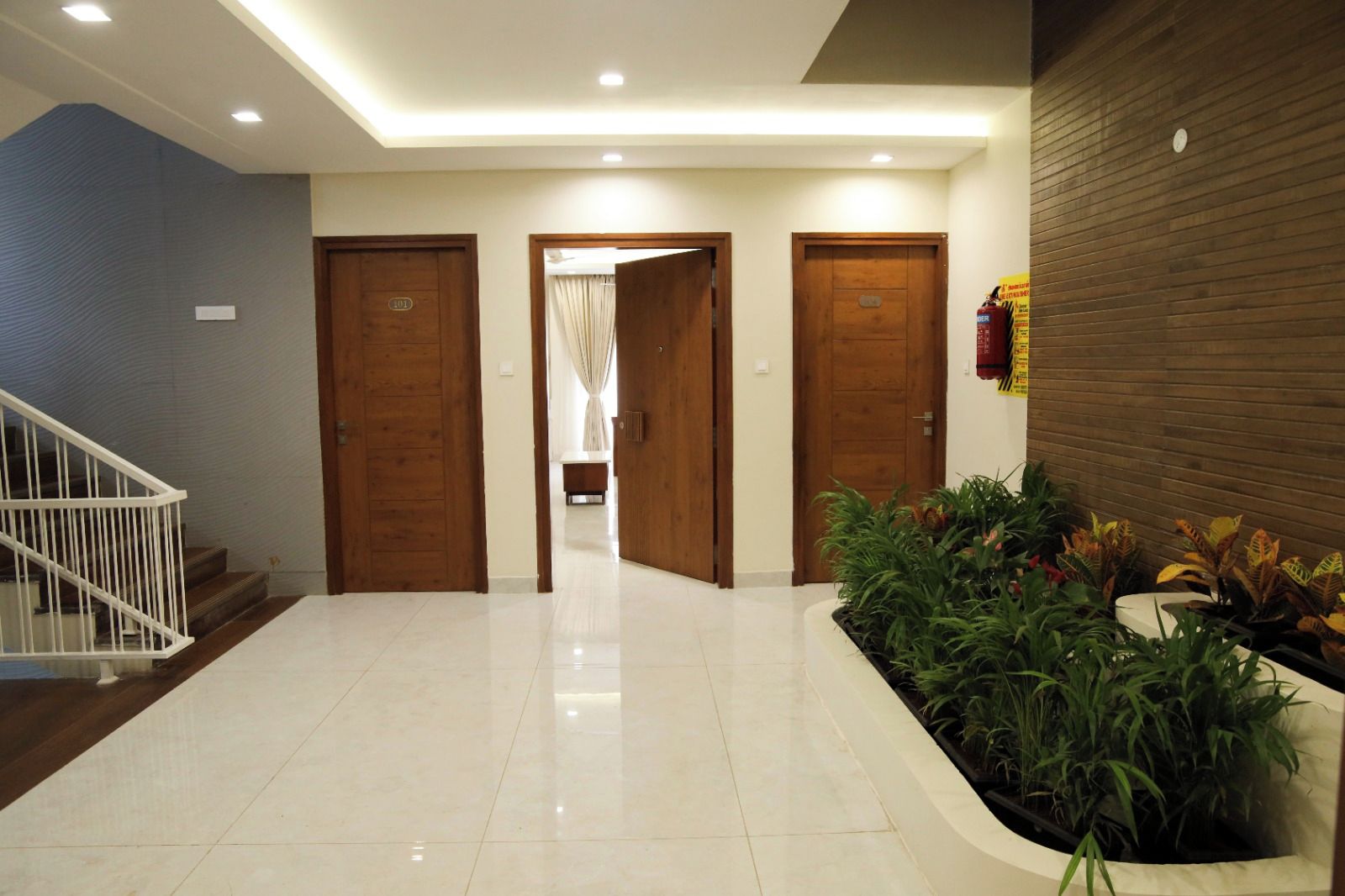 A hotel lobby with a white staircase, three wooden doors, a plant area, and a fire extinguisher on the wall.