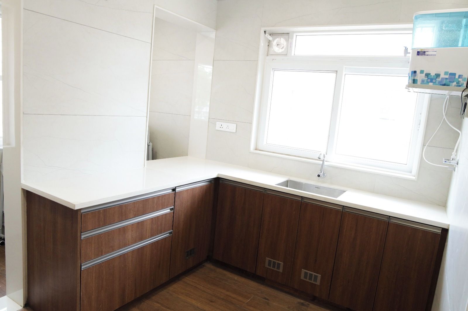 A kitchen with a white countertop, wooden cabinets, a window, and a built-in water dispenser.