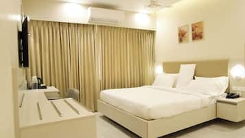 A hotel room with a bed, bedside table, lamp, air conditioning unit, curtains, and artwork on the wall.