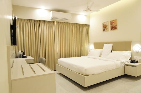 A hotel room with a bed, bedside table, lamp, air conditioning unit, curtains, and artwork on the wall.