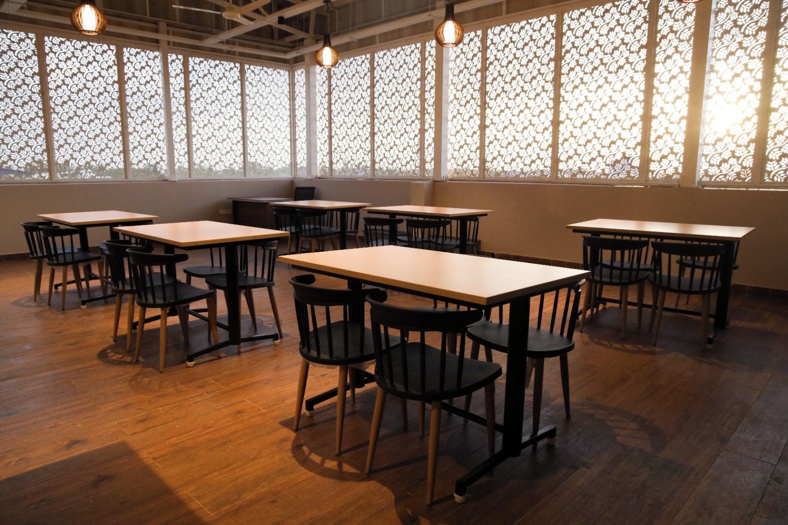 A room with wooden tables and chairs, large windows with patterned curtains, and hanging light fixtures.