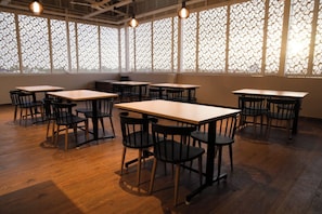 A room with wooden tables and chairs, large windows with patterned curtains, and hanging light fixtures.
