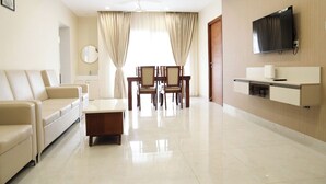 A modern living room with a sofa, a coffee table, a dining table with chairs, a flat-screen TV mounted on the wall, and a door leading to another room. - SS Grandur  (Tiruchirappalli)