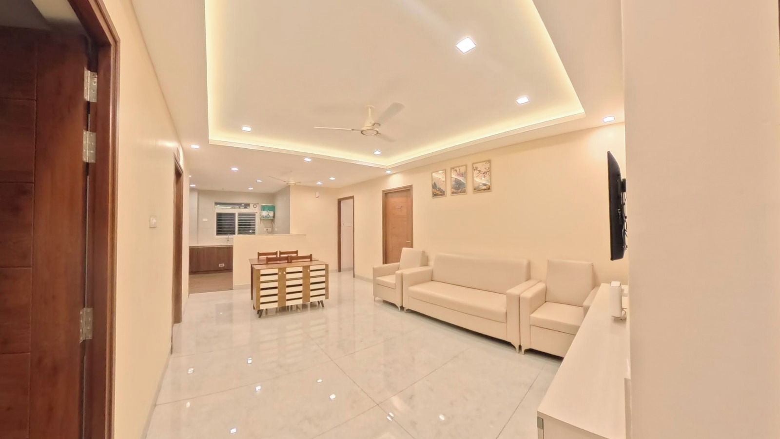 A modern living room with a sofa, a dining table with chairs, a ceiling fan, recessed lighting, and a TV mounted on the wall.