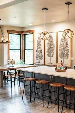 A modern kitchen with a central island, bar stools, pendant lighting, and framed botanical prints on the wall. - Welcome to Seventeen Pines, a stunning Suncadia retreat overlooking Prospector Golf Course’s 17th hole. Enjoy soaring ceilings, a dramatic stone fireplace, private hot tub and beautiful views, all tucked among towering evergreens. (Cle Elum)