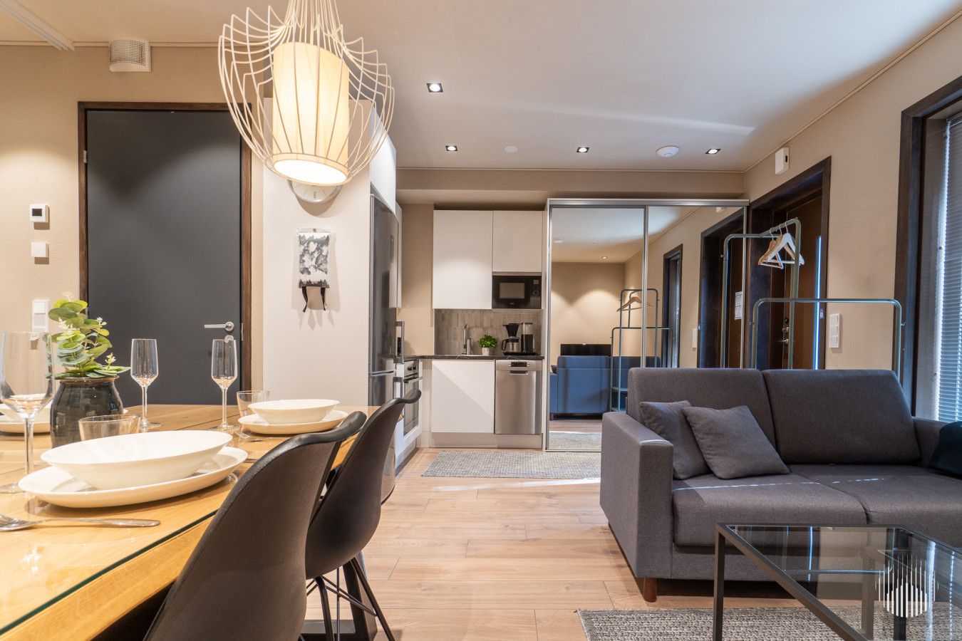 A modern living room with a glass coffee table, a grey sofa, and a dining area with a wooden table and chairs.