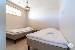 A small, well-lit room with two single beds, a bedside table with a lamp, and a window providing natural light.