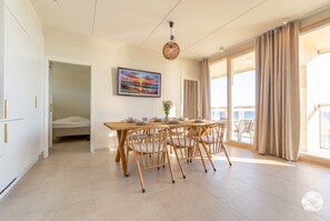 Ein geräumiges Zimmer mit Esstisch, Stühlen, einem Bett und einem großen Fenster mit Vorhängen.