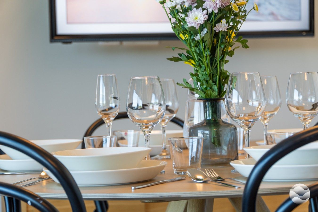Une table à manger dressée avec de l’assiette blanche, des verres à vin transparents et un vase rempli de fleurs.