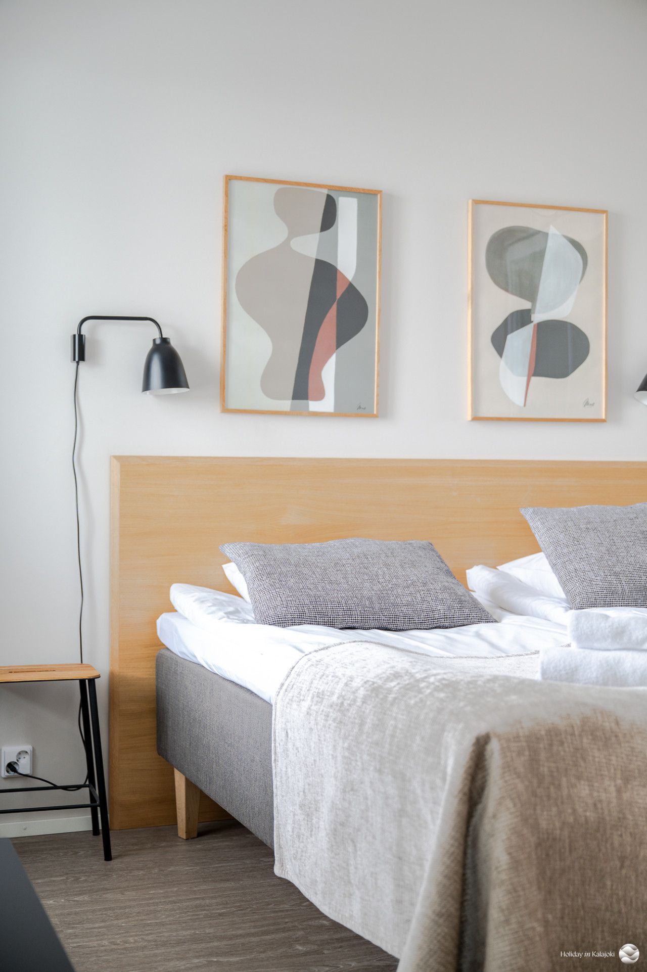 A hotel room with a wooden headboard, a bed with grey bedding, two abstract wall art pieces, a black desk lamp, and a small wooden side table.