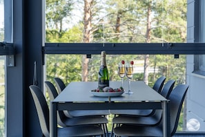 A table with a bottle and two glasses, a bowl of fruit, and a view of trees through a window.