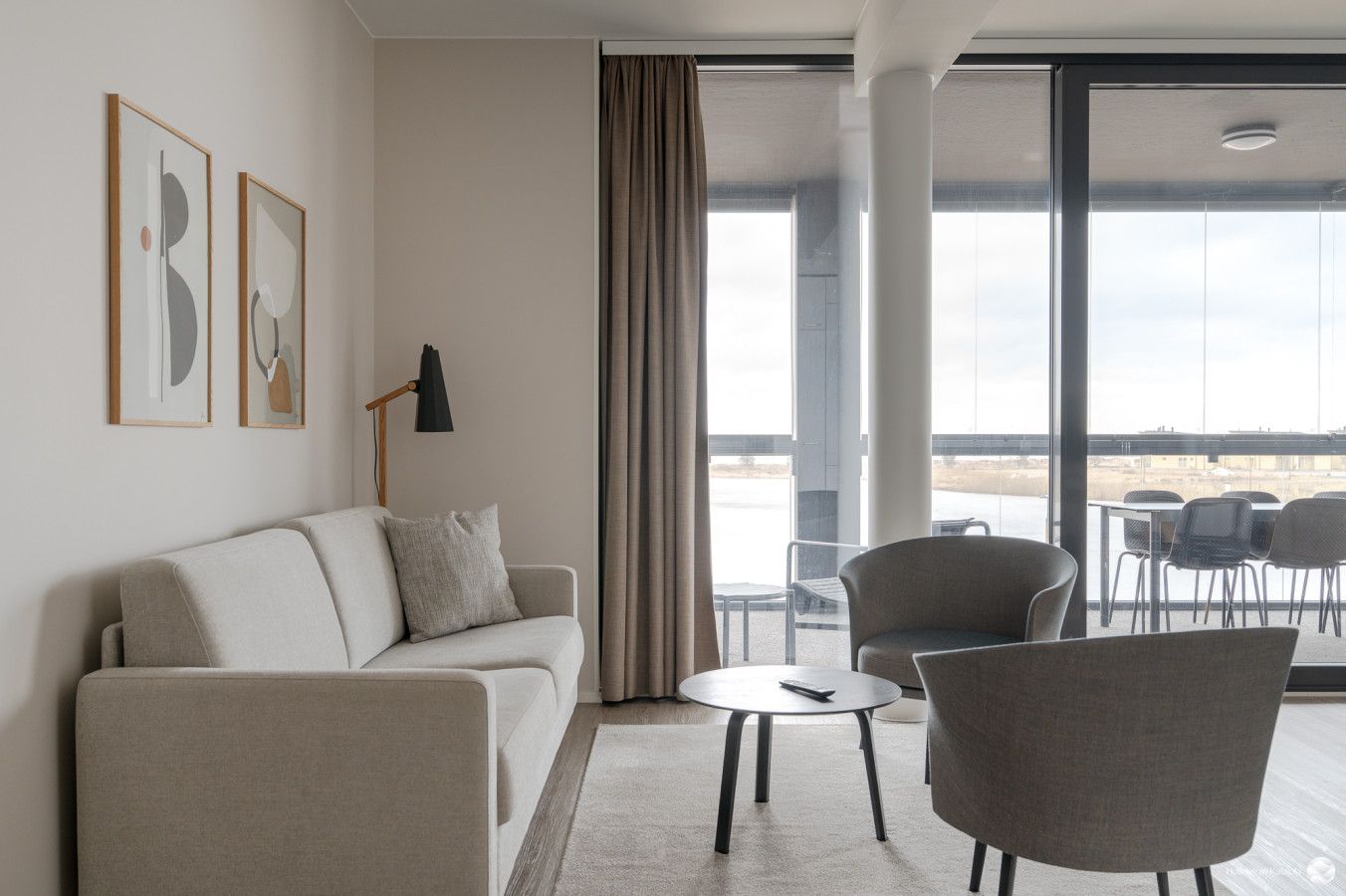 Ein modernes Wohnzimmer mit einer Couch, einem Sessel und einem kleinen Tisch. An den Wänden hängen zwei gerahmte Kunstwerke, und eine Stehlampe sorgt für Licht. Große Fenster bieten einen Blick nach draußen.