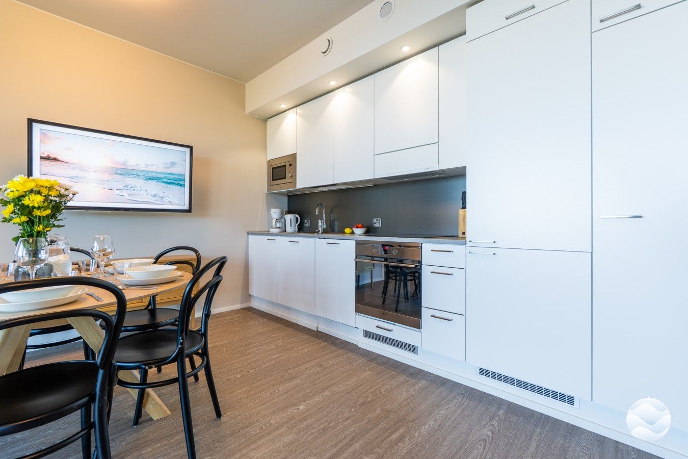 Una cocina moderna con armarios blancos, un comedor con una mesa para dos personas y un cuadro en la pared.