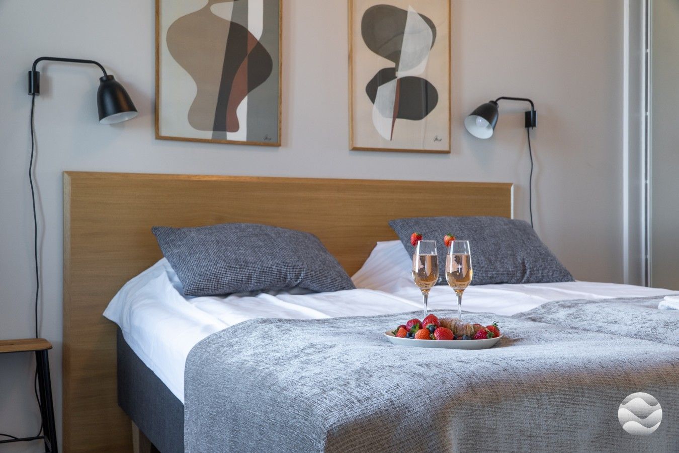 Een net opgemaakt bed met een grijze deken, twee kussens, en een schaal met aardbeien en champagneglazen op het bed.