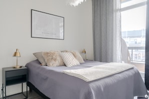 Ein modernes Schlafzimmer mit einem Bett, zwei Nachttischlampen, einer gerahmten Karte und Blick auf die Stadt durch ein Fenster.