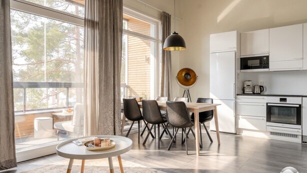 Ett modernt kök med vita skåp, en mikrovågsugn och en kaffebryggare. Här finns också en matplats med ett bord och stolar, samt ett stort fönster med gardiner.