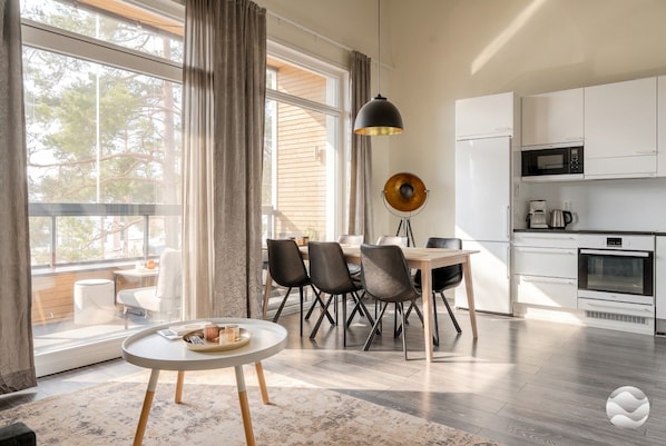 Eine moderne Küche mit weißen Schränken, einer Mikrowelle und einer Kaffeemaschine. Es gibt einen Essbereich mit Tisch und Stühlen sowie ein großes Fenster mit Vorhängen.