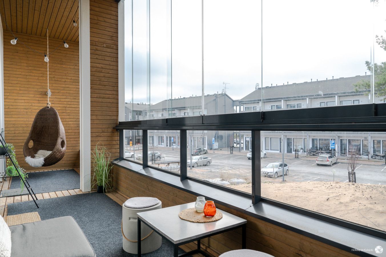 Un intérieur moderne avec un fauteuil suspendu, une table avec une bougie et une vue sur un parking, visible à travers de grandes fenêtres.