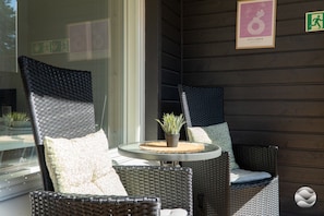 A wicker chair and table set on a balcony with a potted plant, framed pictures on the wall, and an emergency exit sign.