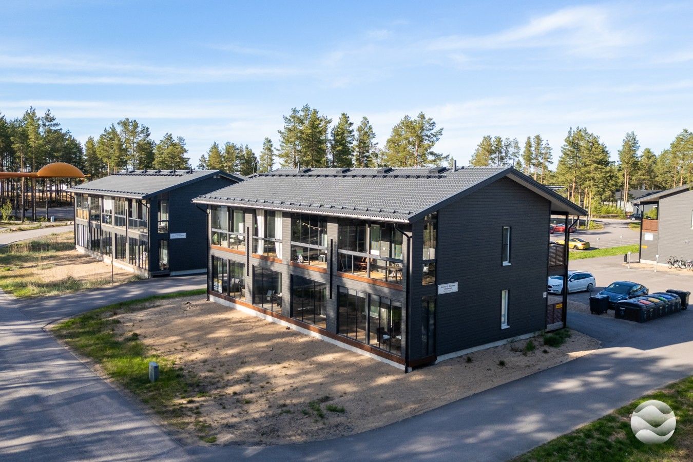 Moderni rakennus, jossa on suuret ikkunat ja tasakatto, ja sen ympärillä on puita ja parkkeerattuja autoja.