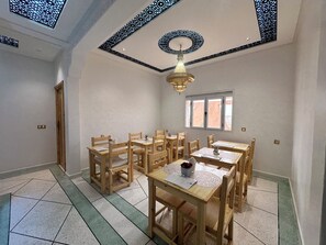 A dining area with wooden tables and chairs, a chandelier, and a window with a view. - Riad Jamilas - Room with Pool Access, Spa & Hammam near Ouarzazate (Ouarzazate)