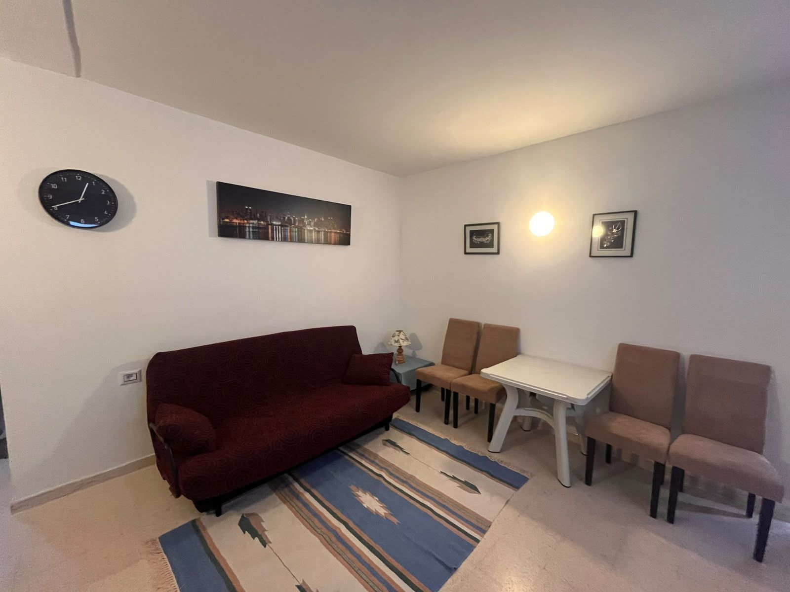 A living room with a red sofa, a small table, two chairs, a clock, and a cityscape painting.