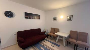 A living room with a red sofa, a small table, two chairs, a clock, and a cityscape painting.