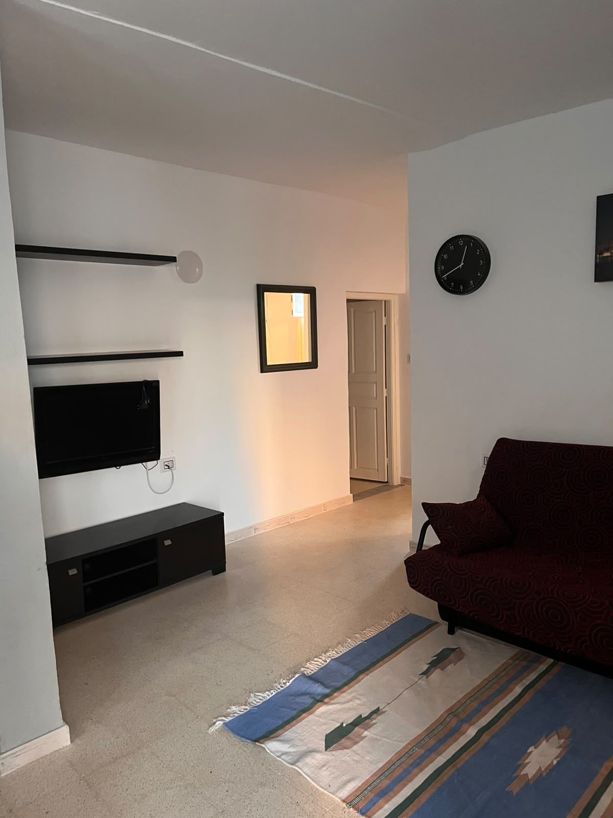 A living room with a flat-screen TV, a red sofa, a patterned rug, and a clock on the wall.