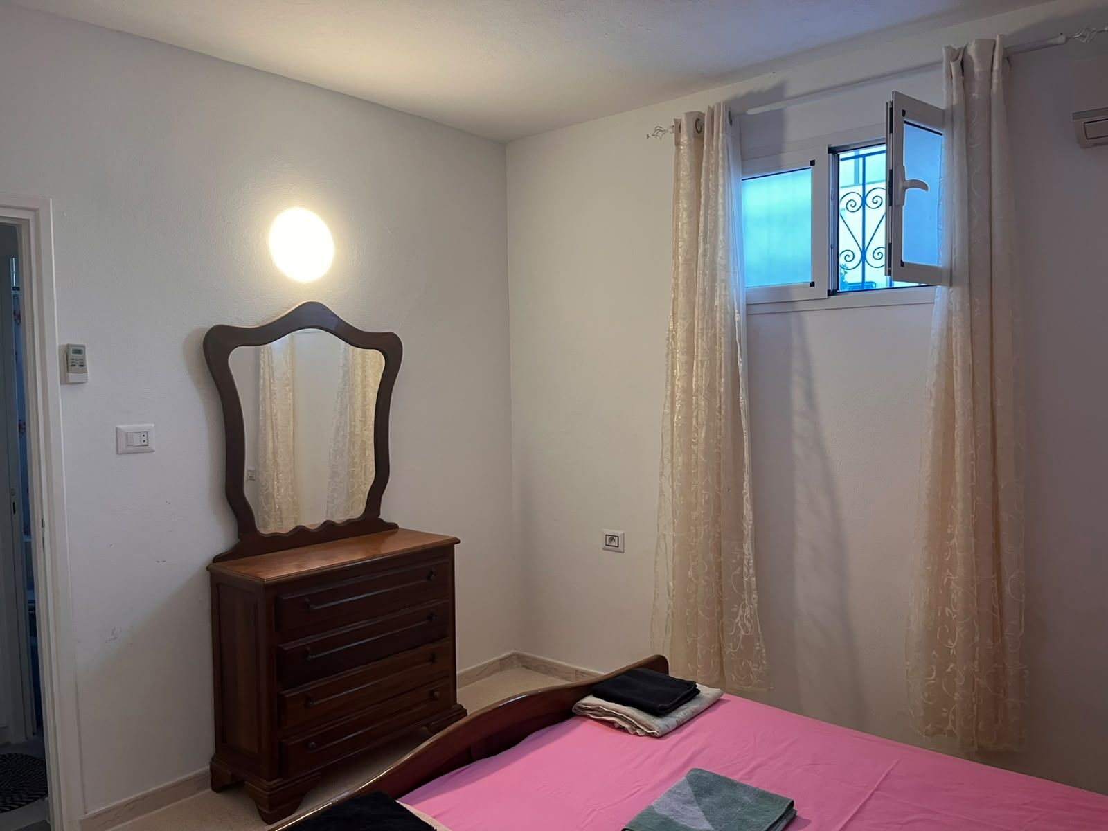 A bedroom with a bed, a dresser, a mirror, a window with curtains, and a wall-mounted light.